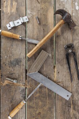 Some carpenter tools on a wooden table. There is a hammer, a pair of pliers, two chisels, a square and screws