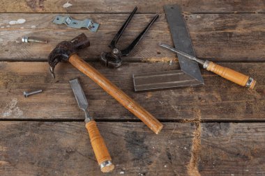 Some carpenter tools on a wooden table. There is a hammer, a pair of pliers, two chisels, a square and screws