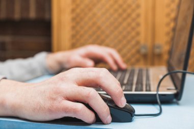 The hands of an unknown person using a laptop, moving the cursor with the mouse and typing with the keyboard, on a blue table.