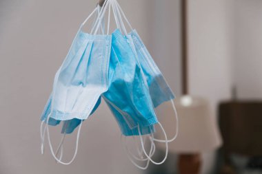 Birçok cerrahi maske kumaş şeritli lastik bantlarla bağlanmış. Arka planda, bir lambanın ayırt edilebildiği bir evin içi odak dışı görünür..