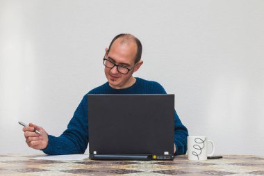 Mavi kazak giymiş, dizüstü bilgisayarın önünde içen gözlüklü bir adam. Masanın üzerinde beyaz bir kupa, bir cep telefonu ve bir sayfa kağıt var. Arka planda beyaz bir duvar var.