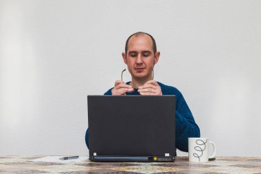 Mavi kazaklı bir adam dizüstü bilgisayarın önünde gözlüklerine bakıyor. Masanın üzerinde beyaz bir fincan, bir akıllı telefon, bir sayfa kağıt ve bir kalem var..