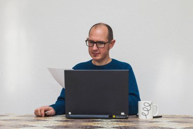 Gözlüklü ve mavi kazaklı bir adam diğer eliyle kalem tutarken dizüstü bilgisayarın önündeki bir kağıt parçasına bakıyor. Masanın üzerinde beyaz bir fincan ve bir akıllı telefon var..