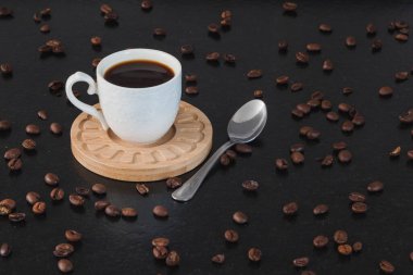 Tahta tabakta kahve, siyah bir granit bankta metal kaşık ve etrafında kahve çekirdekleri olan beyaz bir fincan..