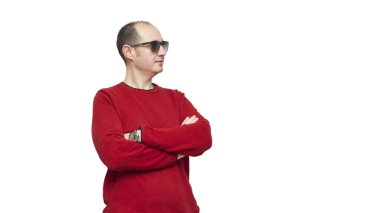 Kırmızı kazak ve güneş gözlüğü takmış kel genç bir adam resmin sağ tarafına bakarak kollarını kavuşturuyor. Arkaplan beyaz.