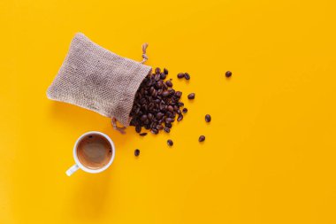 Küçük bir çuval açılır ve kahve çekirdekleri yukarıdan görülebilen sarı bir zemine düşer ve dağılır. Altında kahve dolu beyaz bir kupa var..