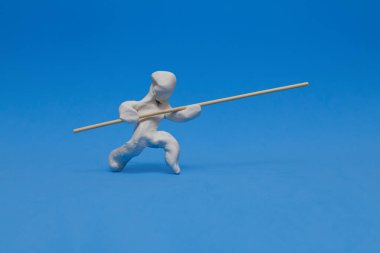 Beyaz bir plastik kukla mavi arka planda sırıkla atlama çalışması yapıyor. Manken direği kaldırarak koşuyor, atlamak için momentum topluyor..