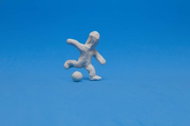 Beyaz plastik bebek mavi arka planda futbol antrenmanı yapıyor. Bebek sağ bacağıyla topa sert bir tekme atmaya hazır..