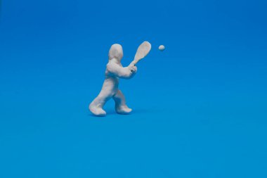 Beyaz plastik bebek mavi arka planda tenis çalışıyor. Oyuncak bebek raketi tutuyor topa vurmak için ona doğru geliyor..