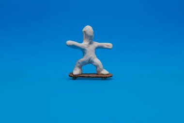 Mavi arka planda kaykay yapan beyaz bir plastik bebek. Bebek yuvarlak kartondan yapılmış kaykayın üzerinde dengede duruyor..