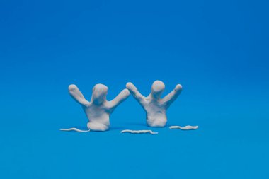Mavi arka planda senkronize yüzme pratiği yapan iki beyaz plastik kukla. Oyuncak bebekler kollarını kaldırmış ve başını plastiklerle çevirmiş bir figür sergiliyorlar..