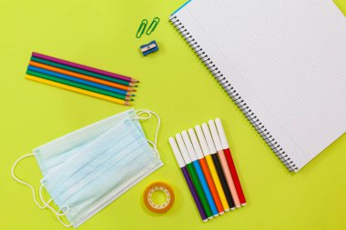 Açık yeşil arka planda iki cerrahi maske, bir defter, iki ataç, bir kalem açacağı, renkli kalemler, renkli kalemler, yapıştırıcı bant ve bir defter var..