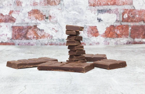 On a gray surface in front of a brick wall is a tower made of ounces of dark chocolate. Around it are more pieces of a chocolate bar.