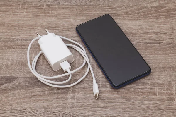 On a wooden table there is a cell phone and a charger with its USB cable. The cable is white and coiled. The smartphone is turned off.