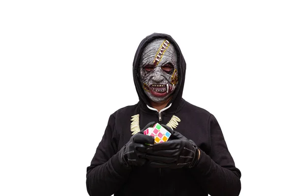 Siyah kapüşonlu sweatshirt giyen zombi maskesi takmış bir adam bir Rubik küpünü çözüyor..