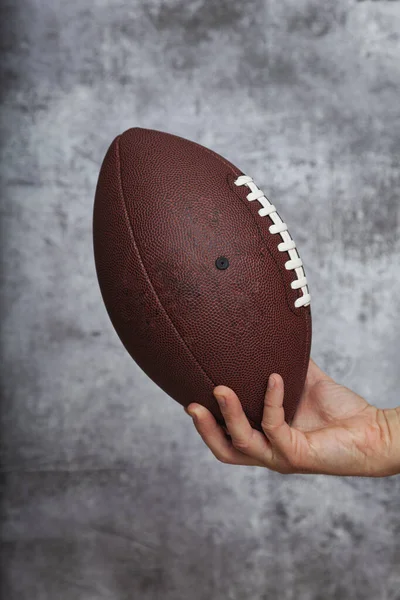 茶色のアメリカンフットボールボールを持っている男の右手テクスチャグレーの背景に白い文字列 — ストック写真