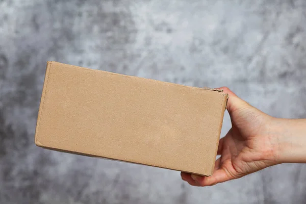 A man\'s right hand holding or delivering a cardboard box that may be a package shipped on a textured gray background.