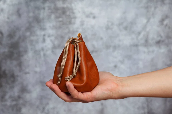 A man\'s right hand holding a brown leather bag closed with a string containing money inside on a textured gray background.