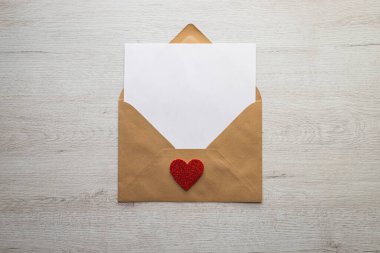 Beyaz ahşap bir masada üzerinde kırmızı bir kalp olan kahverengi bir zarf. Zarf açık ve içindeki kağıt beyaz ve boş..