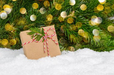 Köknar ağacının altında el yapımı plastik Noel hediyesi. El işi kağıt ve ipe sarılı hediye kutusu karlar içinde duruyor. Arkaplan olarak çam ağacı dalları ve ışık var.