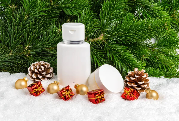 Set of winter skin care cosmetic products in snow under Christmas tree with gifts, pine cones and baubles. Face cream jar and body lotion bottle. Present for holiday and seasonal skin protection