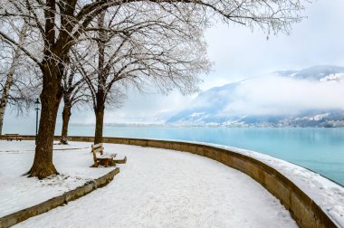 Zell am See in winter. Esplanade along Lake Zell, snow, frozen trees and misty mountain in alpine town. Famous ski resort in Alps mountains, Austria. Winter wonderland holidays destination