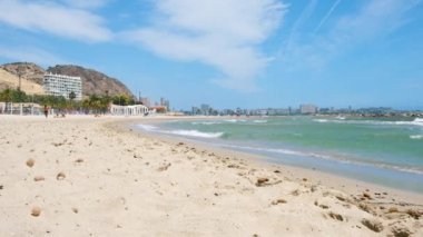 ALICANTE, İspanya - 17 Mayıs 2021: Alicante 'de denizde yürüyen ve yüzen turistlerle Playa del Postiguet manzarası. Dalgalar arka planda kumlu sahile, dağa ve kasabaya çarpıyor.