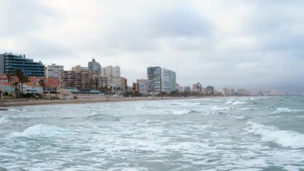 Gelombang menerjang pantai San Juan berpasir di Alicante, Spanyol pada hari berawan. Costa Blanca wilayah di laut Mediterania. Orang-orang tak dikenal berjalan di pantai — Stok Video
