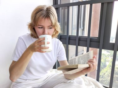 Pencerenin kenarında oturan, kitap okuyan ve içecek içen çekici sarışın kadın.