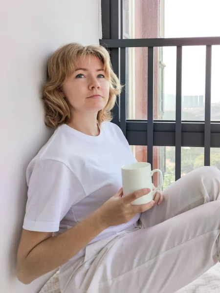 Atractiva joven sentada en el alféizar de la ventana y sosteniendo una taza Fotos de stock libres de derechos