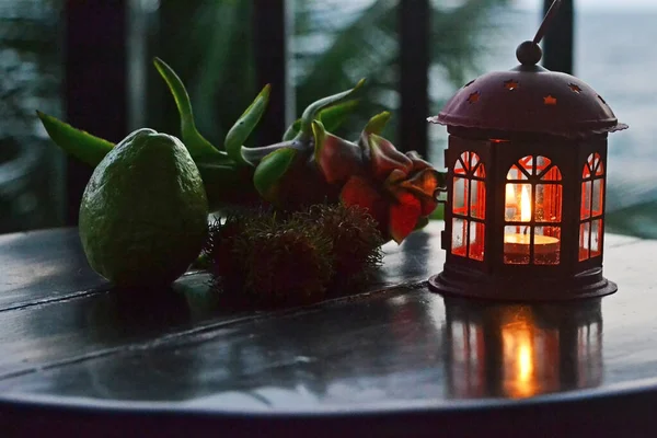 半円形の窓のある家の形をした燭台の小さなろうそくは 丸い木製のテーブルの上にあり 隣にはトロピカルフルーツがあります — ストック写真