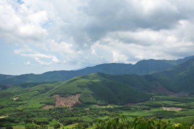 Güneşin aydınlattığı dağlar ve vadi manzarası, bulutların altında, bir sürü ağaç ve yeşillik.