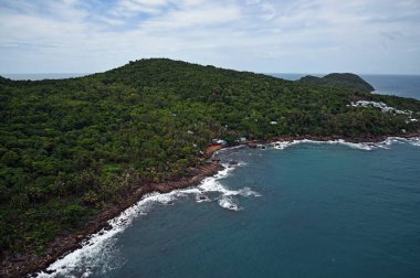 Deniz, kıyı şeridi ve adanın sonu, tepeler, taşlar, orman ve balıkçı evleri, üst manzara, beyaz deniz köpüğü.