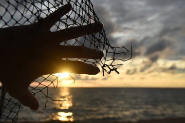 Bir el, deniz batımının arka planında yırtık bir kenarla bir halat ağın üzerinde uzanır.