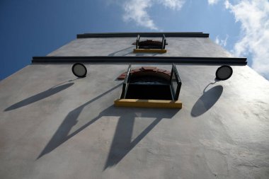 Güneşli bir günde iki penceresi açık, iki lambası gölgeli alçılı iki katlı bir evin alt manzarası.