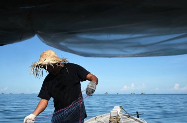 Vietnam, Phu Quoc - 27 Haziran 2021. Siyah tişörtlü ve hasır şapkalı bir balıkçı teknenin pruvasında duran ağları denizden çeker. 