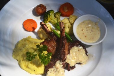 Soslu ızgara kaburga, patates püresi ve sebzeler beyaz bir tabakta, güneşle aydınlatılmış, üst manzara