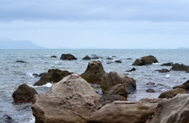 Kayalık sahilden denize, ufukta adalar, ön planda büyük taşlar.