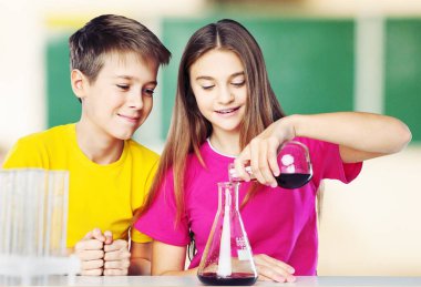 Küçük bir kız renkli bir sıvının üzerine dökerken küçük çocuğun matarayı sabit tuttuğu bir kimya deneyi yapıyor.