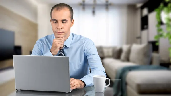 一个心事重重的年轻人坐在家里的桌子旁 一边上网一边用笔记本电脑 — 图库照片