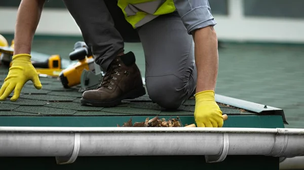 A man worker is cleaning a clogged roof gutter from dirt, debris and fallen leaves to prevent water and let rainwater drain properly.