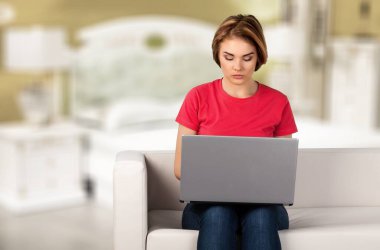 Gülümseyen genç bir kadın, evden çalışıyor, kanepede oturuyor, dizüstü bilgisayar kullanıyor.,