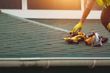 Koruma işinde çalışan çatı tamircisi eldiven giyer, inşaat binasının altındaki yeni çatının tepesine elektrikli alet kullanır veya inşa eder.