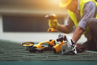 Koruma işinde çalışan çatı tamircisi eldiven giyer, inşaat binasının altındaki yeni çatının tepesine elektrikli alet kullanır veya inşa eder.