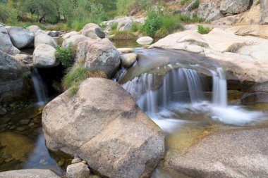 Temiz bir dağ deresinde pürüzsüz granit kayalar arasında akan küçük bir şelalenin yakın görüntüsü. Sahne, Sierra de Gata bölgesinin doğal güzelliğini, tatlı su, yosun ve yeşillik ile sakin ve barışçıl bir atmosfer yaratarak yakalar. Kimlik