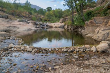 Temiz bir dağ deresinde pürüzsüz granit kayalar arasında akan küçük bir şelalenin yakın görüntüsü. Sahne, Sierra de Gata bölgesinin doğal güzelliğini, tatlı su, yosun ve yeşillik ile sakin ve barışçıl bir atmosfer yaratarak yakalar. Kimlik