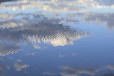 Dinamik bir su yüzeyinin, parlak mavi gökyüzünü ve dramatik beyaz ve gri bulutları yansıtan yakın plan, soyut bir görüntüsü. Sudaki ince dalgalanmalar yansımayı çarpıcı bir yatay dalga desenine ve dokularına dönüştürür.