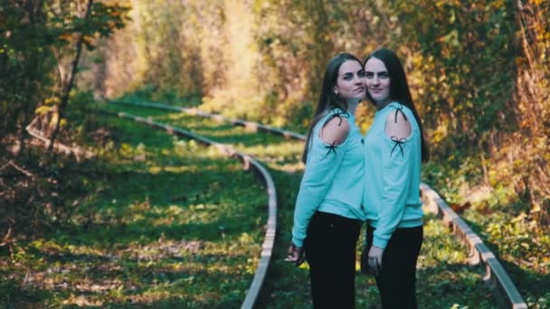 Sorrindo jovens gêmeas mulheres de pé juntos na estrada de ferro. Olhe para a câmera ao ar livre. — Vídeo de Stock