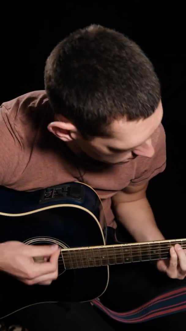 Vidéo verticale. Young Man Blogger joue de la guitare acoustique sur fond noir — Video