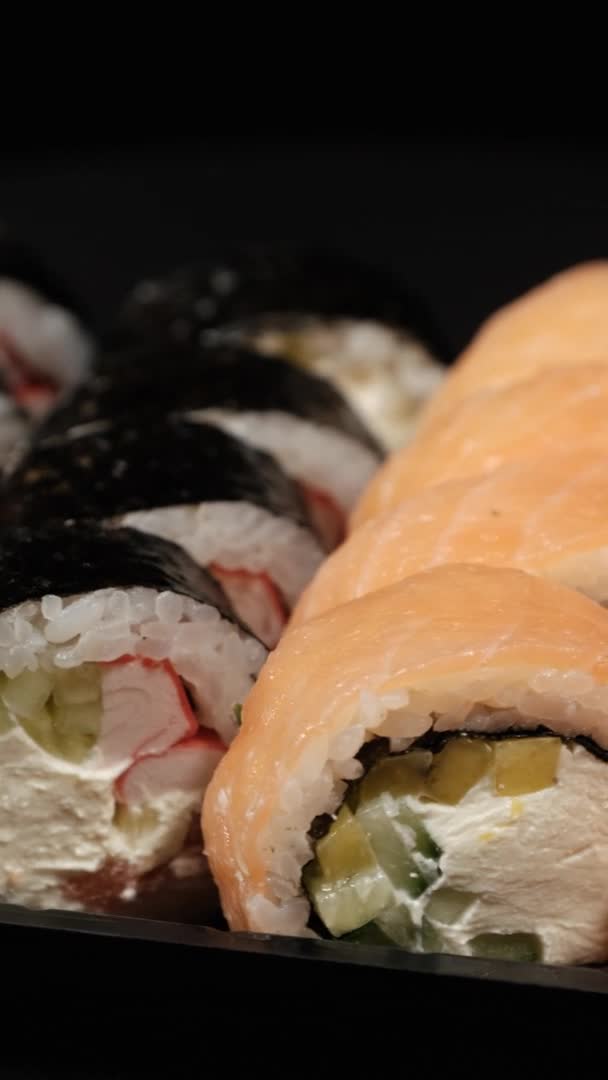 Vídeo vertical. Rollos de sushi gira en un contenedor de plástico sobre un fondo negro — Vídeo de stock